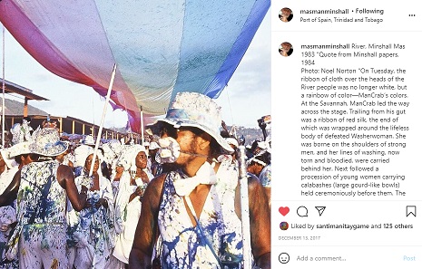 Masqueraders play mas under a rainbow banner spanning the entire band, wearing white splattered with messy muddy colours