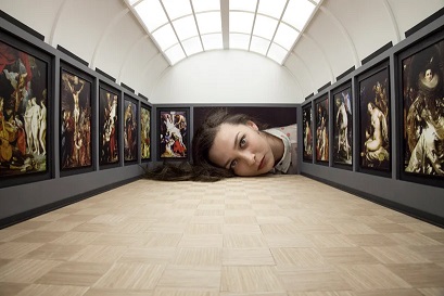 gallery of fine classical art interrupted by a woman's head that takes up almost an entire wall. She is on the wall like the art, larger than life in this miniature gallery