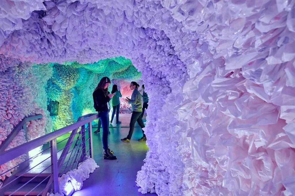 people walk through a pastel coloured space where the walls are thick with plastic bags that pick up the colours in the sapce and create an almost cloud-like softness to the space