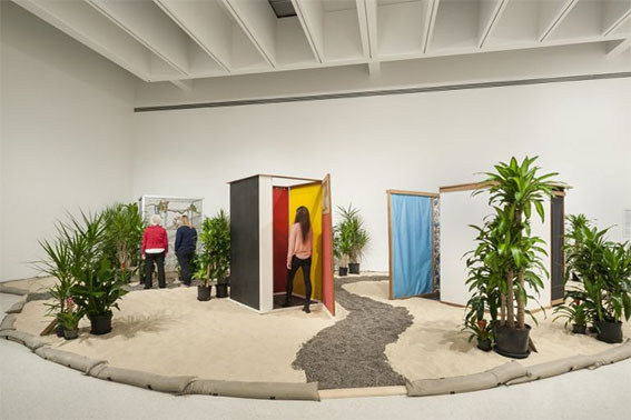art installation showing a large mound of sand with tall potted plants scattered about, and gravel pathways leading to little shacks made of plywood and fabric.