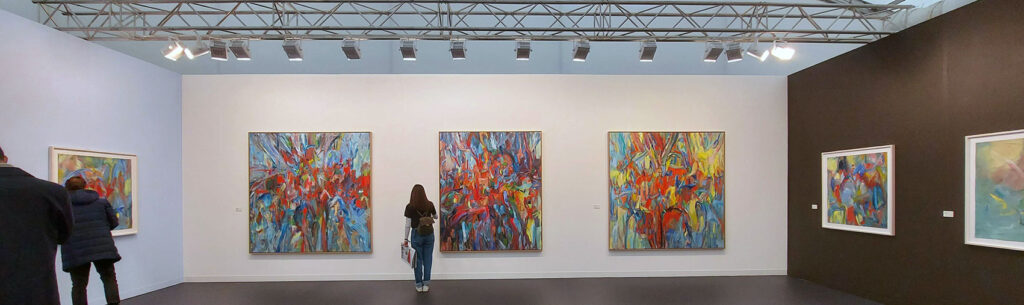 3 large painting with a glorious mess of abstract colour against a white wall with a rail of lighting overhead and a woman watching them