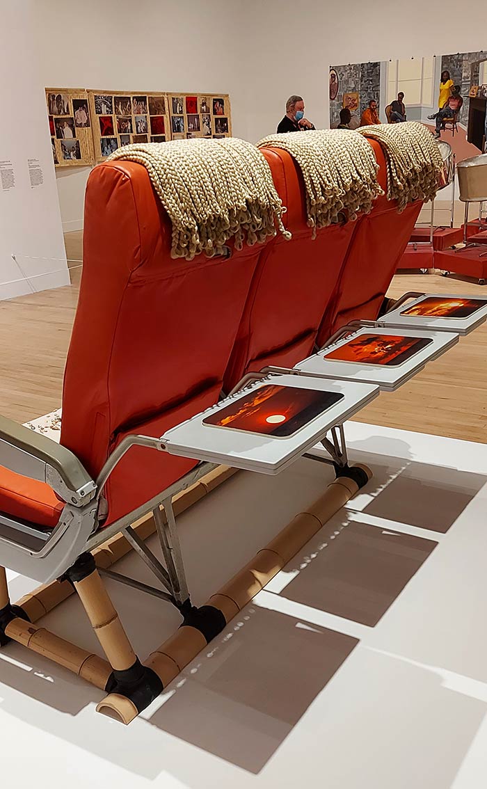 rearview of 3 red-orange airline seats with airline trays down, each with a sunset pictured on it. The row of 3 seats is anchored on a bamboo frame