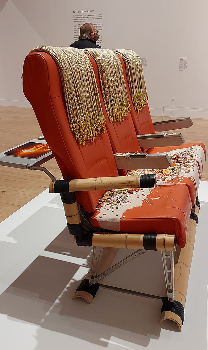 front view of 3 red-orange airline seats with sand, shells and beads on the seats. The row of 3 seats is anchored on a bamboo frame, which supplements a missing armrest