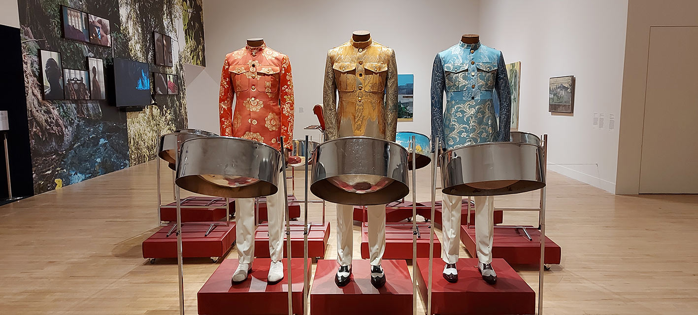 2 headless mannequins dressed in fancy paisley jackets - 1 orange and gold, one gold and silver, one blue and silver. Each is wearing sharply pressed white trousers. 2 mannequins are in 2-toned shoes, all are standing behind on a red pedestal behind a tenor pan each. Several other steelpans with no 'players' also stand on red pedestals behind the three mannequins