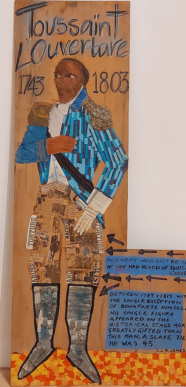 mixed media collage on wood that forms the body of a man in old colonial uniform that features a blue jacket with golden epaulettes, black sash, breeches and tall black boots. The words Toussaint L'Ouverture 1743 - 1803 are drawn above his head
