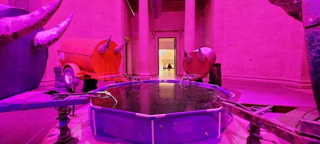 Room bathed in red-pink lighting with a paddling pool full to the brim in the middle. Drinking from or filling up the pool are four cylindrical iron gas tanks perched on stacked tyres, tilted towards the pool with papier-mache horns attached to the 'pool' end of the tank.