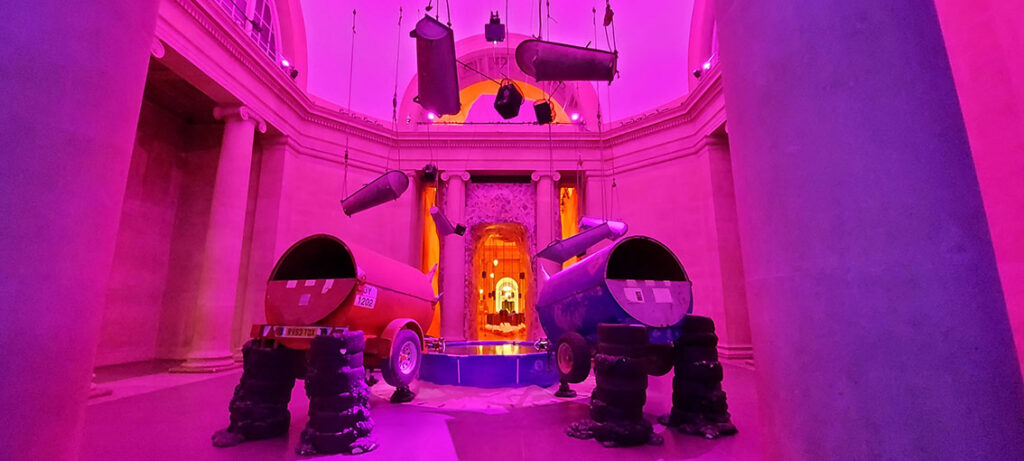 Room bathed in red-pink lighting with a paddling pool full to the brim in the middle. Drinking from or filling up the pool are four cylindrical iron gas tanks perched on stacked tyres, tilted towards the pool with papier-mache horns attached to the 'pool' end of the tank.