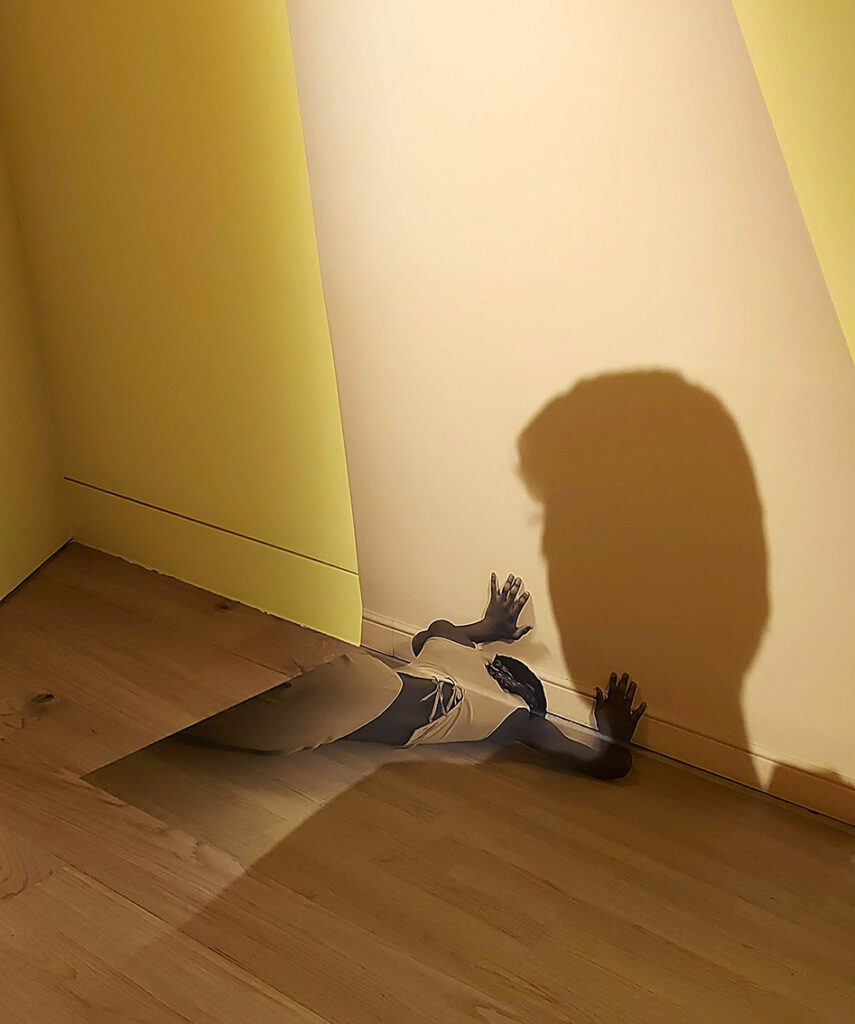 photo of a dancer laying face down on the floor pushing against the wall, positioned so the hands are actually against the wall and gives the illusion of the dancer really being there.