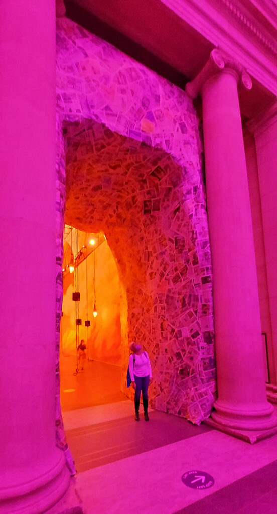 Stacey standing in a giant archway, looking incredibly tiny. The lighting is very pink and the arch is made from papier-mache.