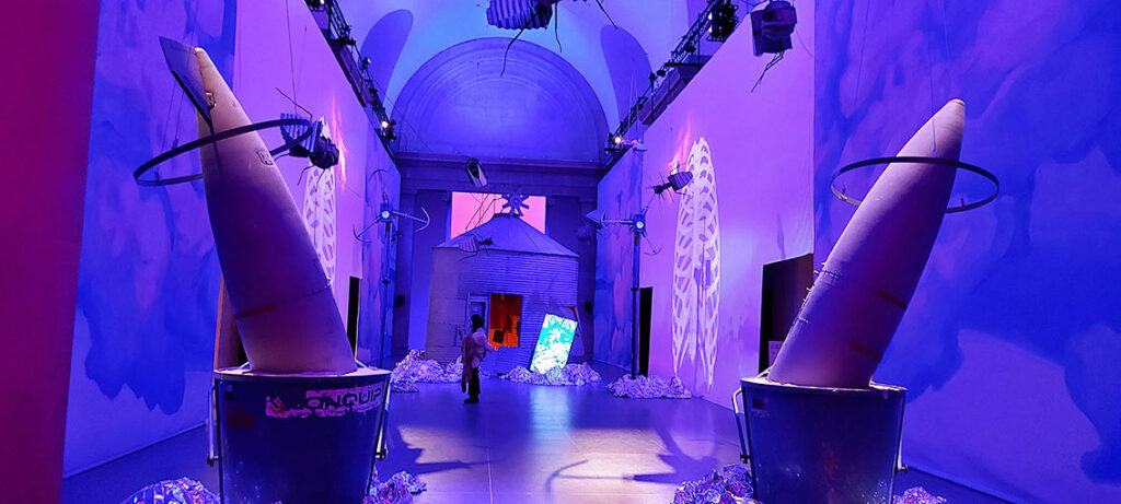 photo of the celestial room as described in text - cloud paintings and projections on either side with the galvanized circular kiln or still like structure in the background. the broken torpedo sunk into iron buckets of sand sporting metal hoop halos is in the foreground